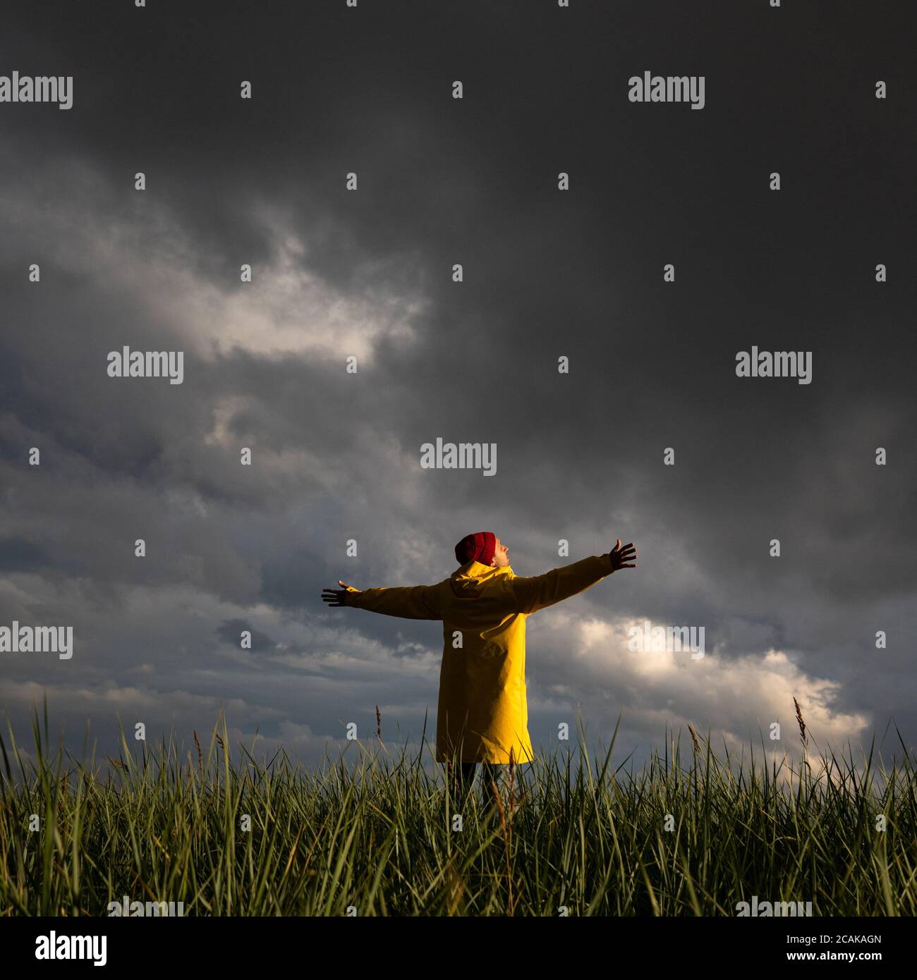 Homme en imperméable jaune et chapeau rouge avec les mains écartant largement ouvertes debout sur le terrain par temps pluvieux, regarde le ciel nuageux dramatique. Saison d'automne. Banque D'Images