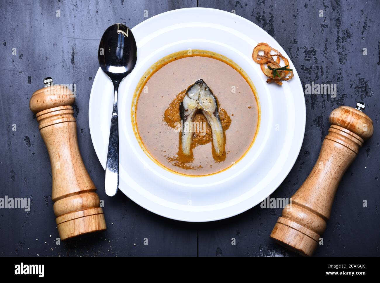 Soupe de poisson avec un morceau de poisson et des rondelles d'oignon frits sur une assiette blanche. Poivrière en bois, saltcave et cuillère près du plat. Concept de cuisine française. Soupe à la crème ou purée brune sur fond de texture grise Banque D'Images