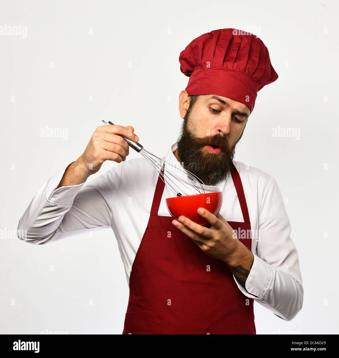Chef mélangeant ou foulant des ingrédients dans une assiette rouge. Cuire  avec un visage curieux en uniforme bordeaux utilise un fouet et un bol.  Homme avec barbe tient les ustensiles de cuisine