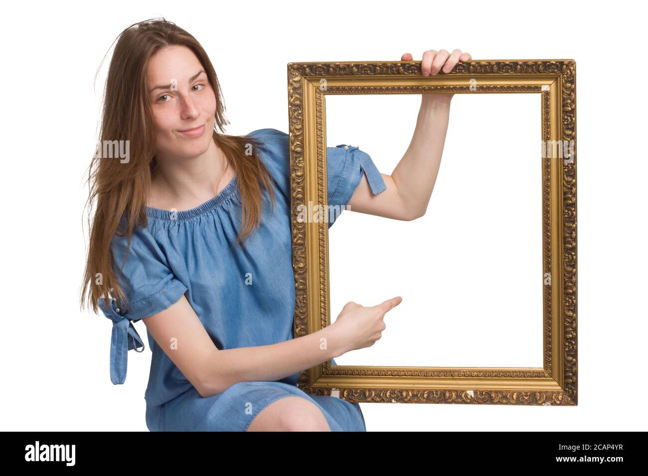 Une jeune fille avec de longs cheveux dans une robe bleue. Isolé sur un fond blanc, tenant un cadre d'image dans ses mains, pointant vers lui avec son doigt. copyspace. Avec un regard malicieux Banque D'Images