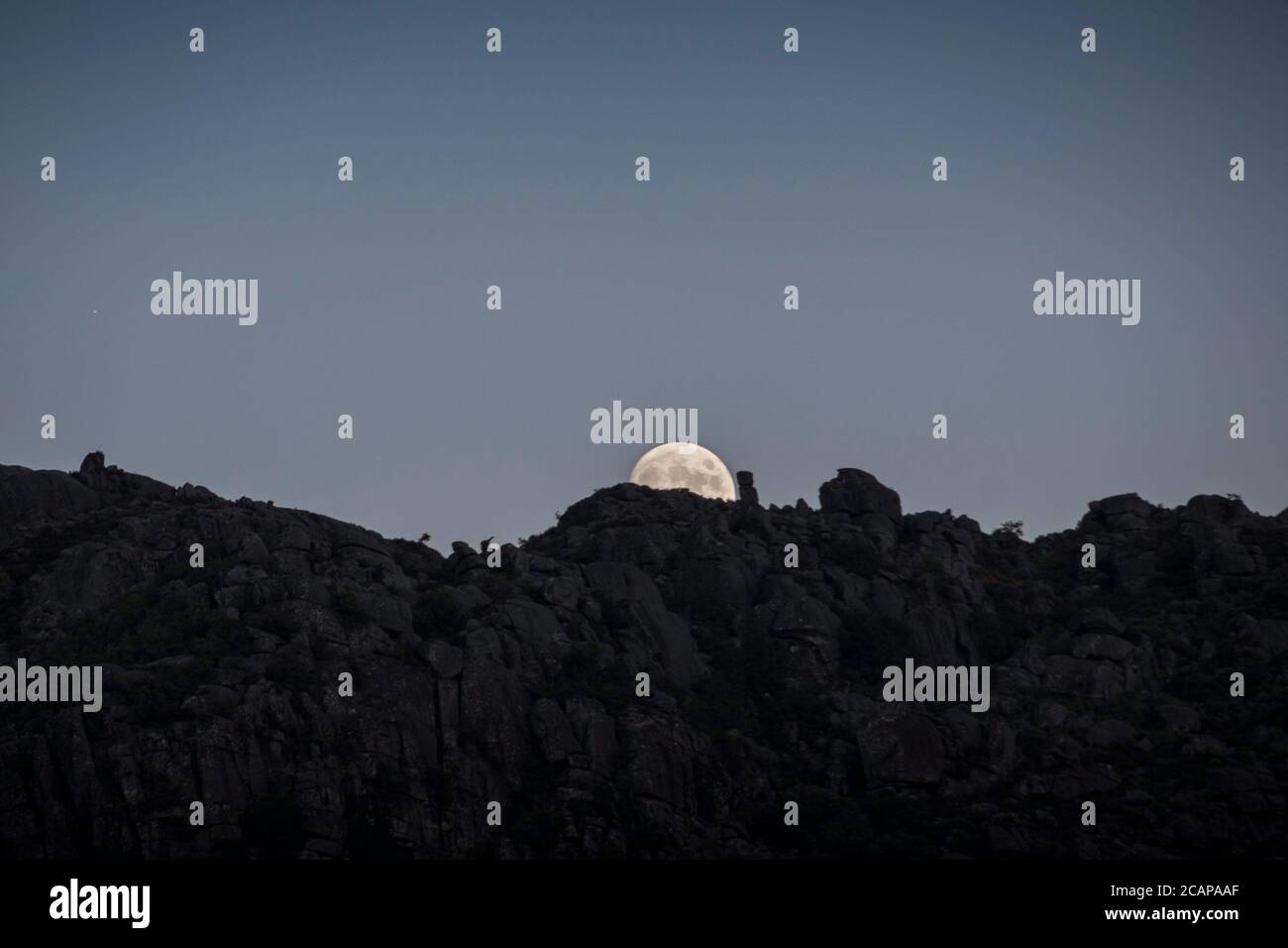 La lune presque complète s'élève lentement derrière des pierres sur les sommets de montagne en un moment de crépuscule, loin dans la distance Banque D'Images