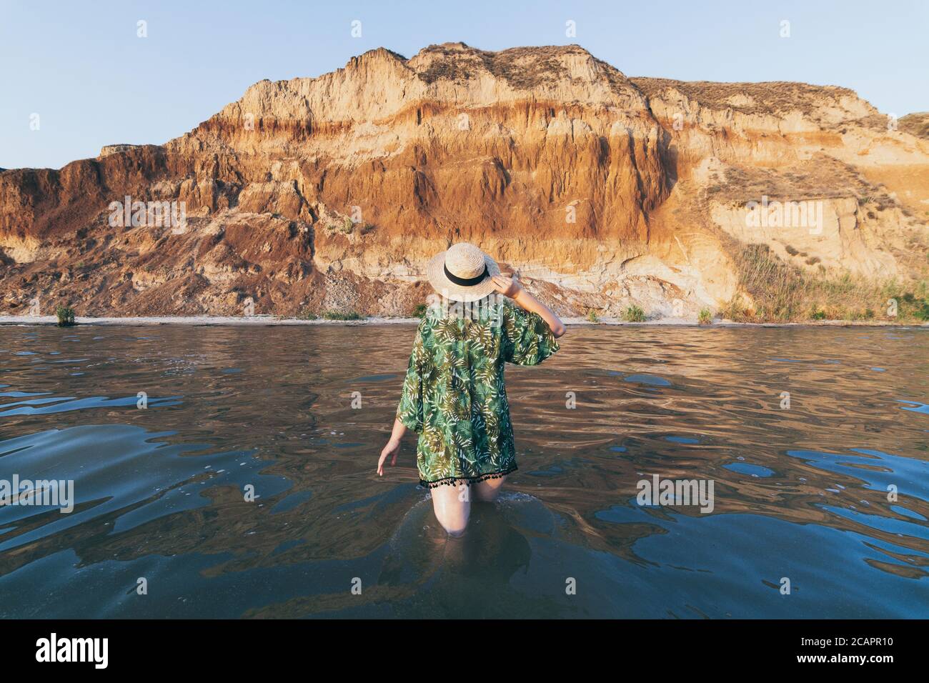 Femme en chapeau de paille marchant dans l'eau de mer donnant sur les montagnes. Banque D'Images