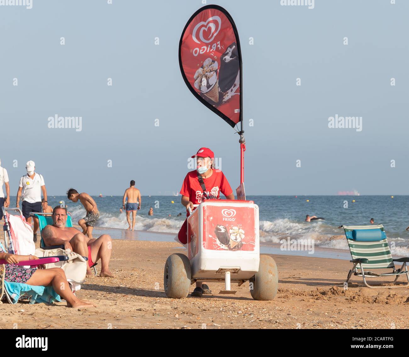 Punta Umbria, Huelva, Espagne - 7 août 2020: Une vendeuse de rue féminine vendant de la glace sur la plage. Le vendeur porte un masque de protection ou un masque médical Banque D'Images