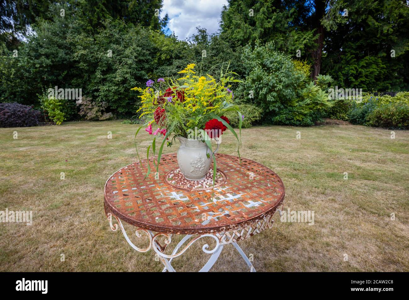 Arrangement de fleurs: Une exposition de fleurs d'été cueillies dans un jardin anglais mélangé disposés dans une cruche de drabware sur une table de jardin en métal d'époque rouillée Banque D'Images