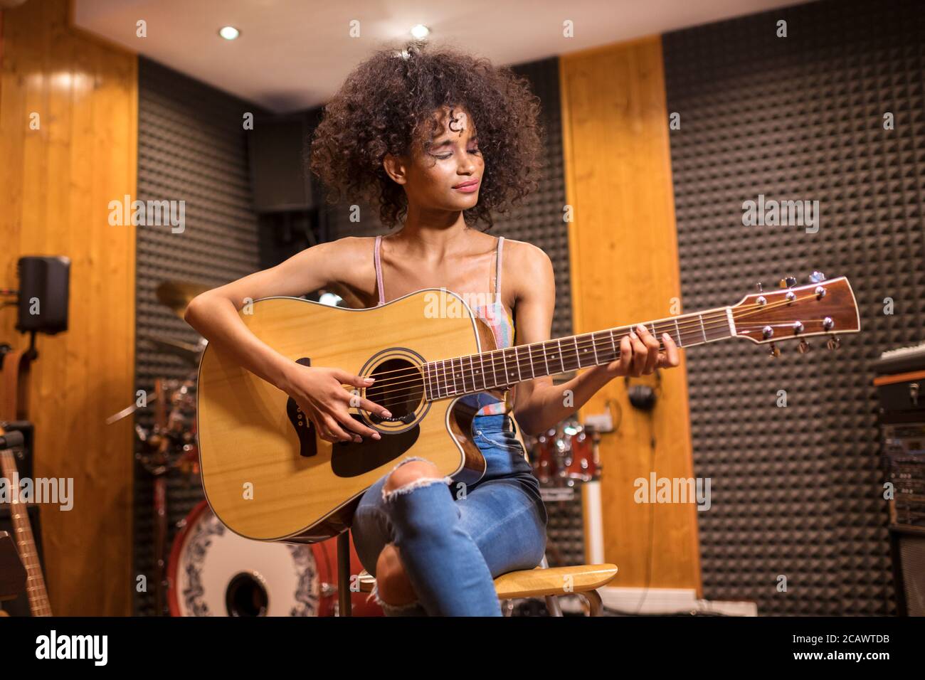 Jeune fille noire à la mode avec les cheveux longs bouclés assis jouant guitare dans un studio d'enregistrement avec un sourire de plaisir en gros plan Banque D'Images