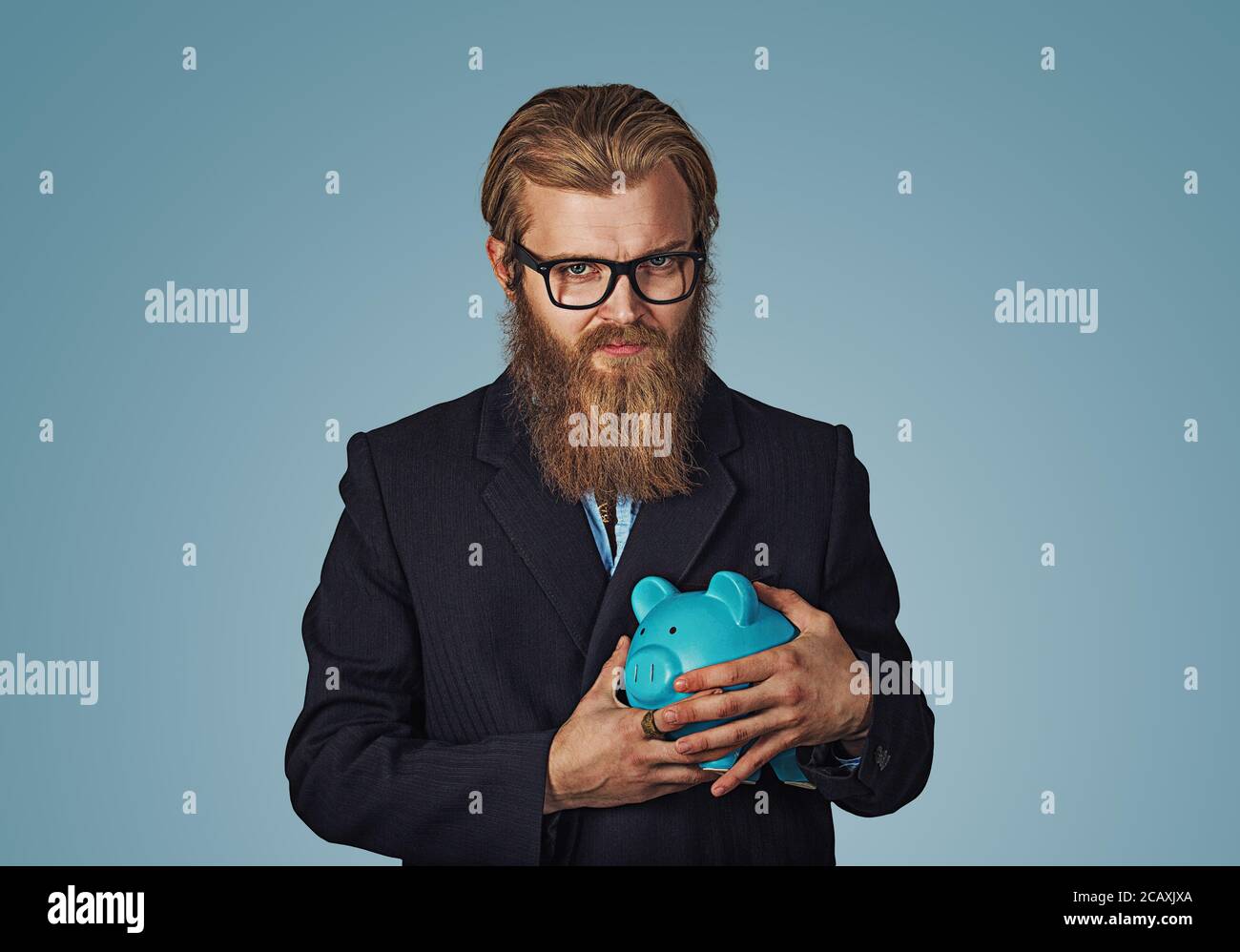Jeune homme d'hipster barbu avare cupide man holding piggy bank isolé sur fond mur studio bleu. Face à l'émotion humaine expression négative Banque D'Images