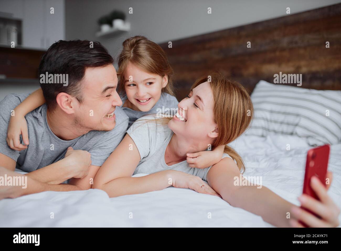jeunes parents caucasiens allongé sur le lit avec une petite fille enfant, utiliser un smartphone rouge et prendre selfie, émotionnellement famille heureuse Banque D'Images