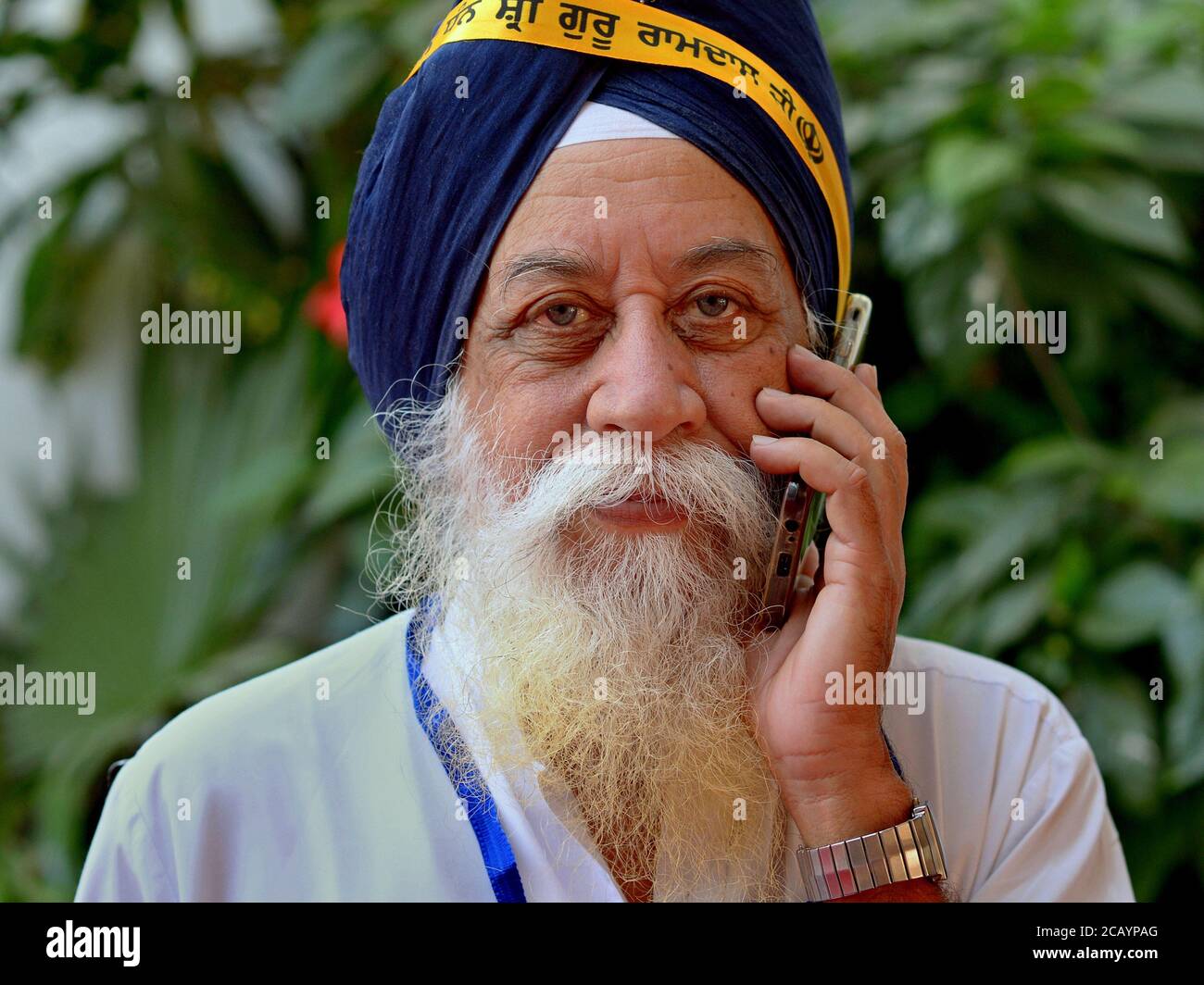 Le vieil Indien Sikh avec turban bleu (dastar) fait un appel téléphonique sur son téléphone cellulaire et regarde dans la caméra. Banque D'Images