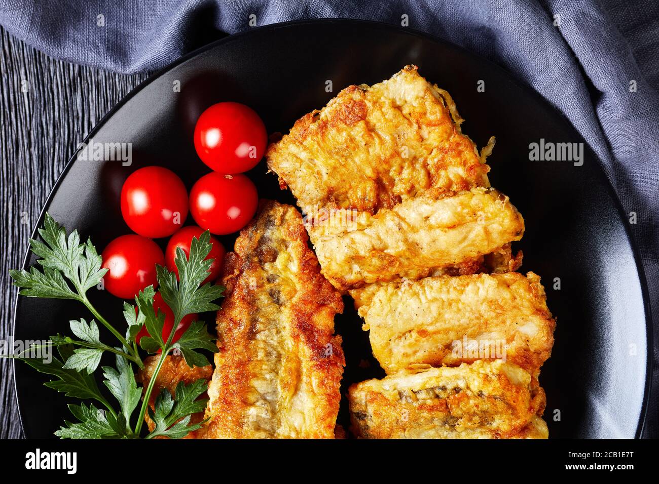 Vue depuis le dessus du filet de cabillaud frit servi sur une assiette noire avec tomates cerises, persil frais sur fond de bois foncé, gros plan, plat Banque D'Images