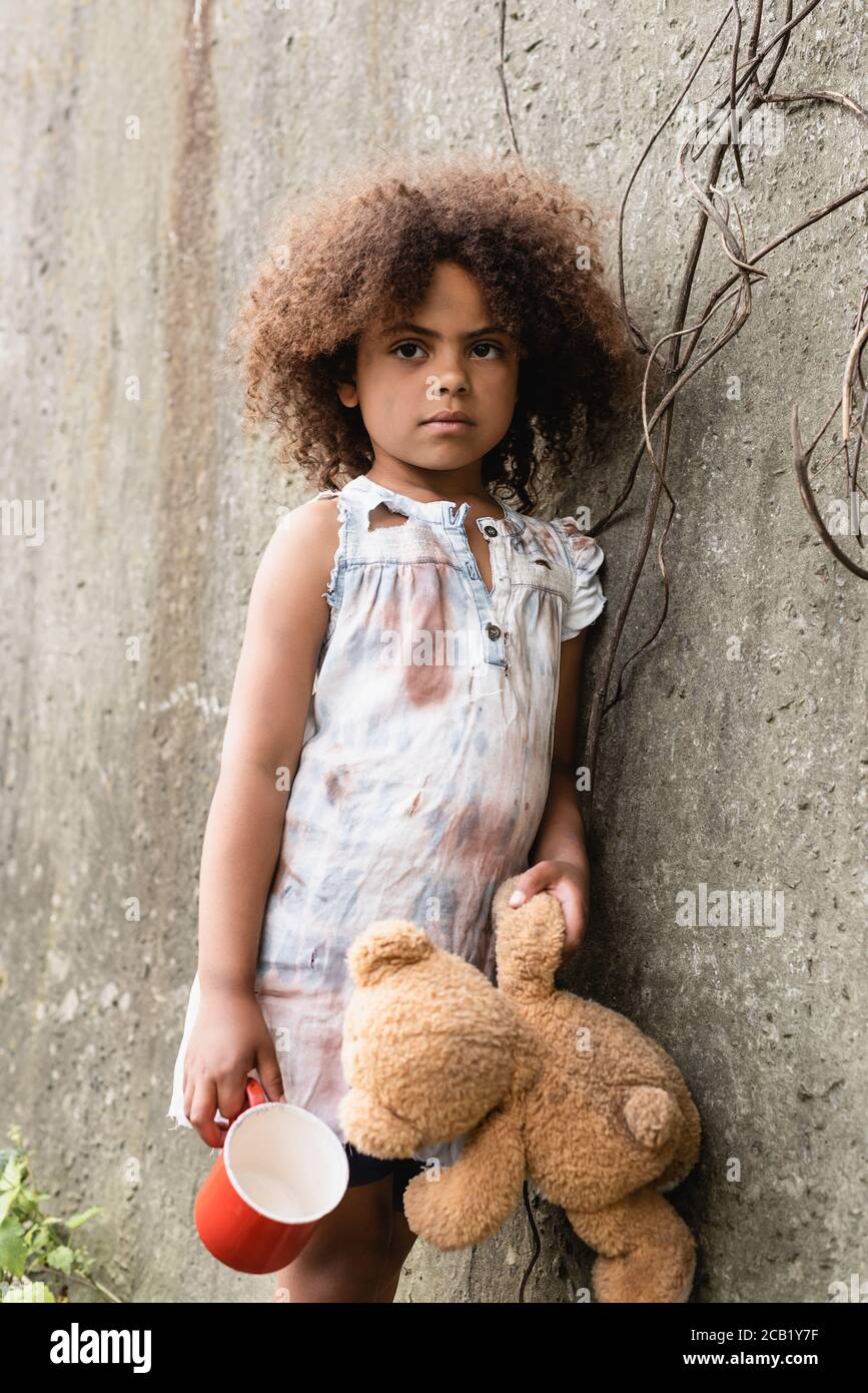 Mendiant enfant afro-américain tenant l'ours en peluche et la tasse près mur en béton sur la rue urbaine Banque D'Images