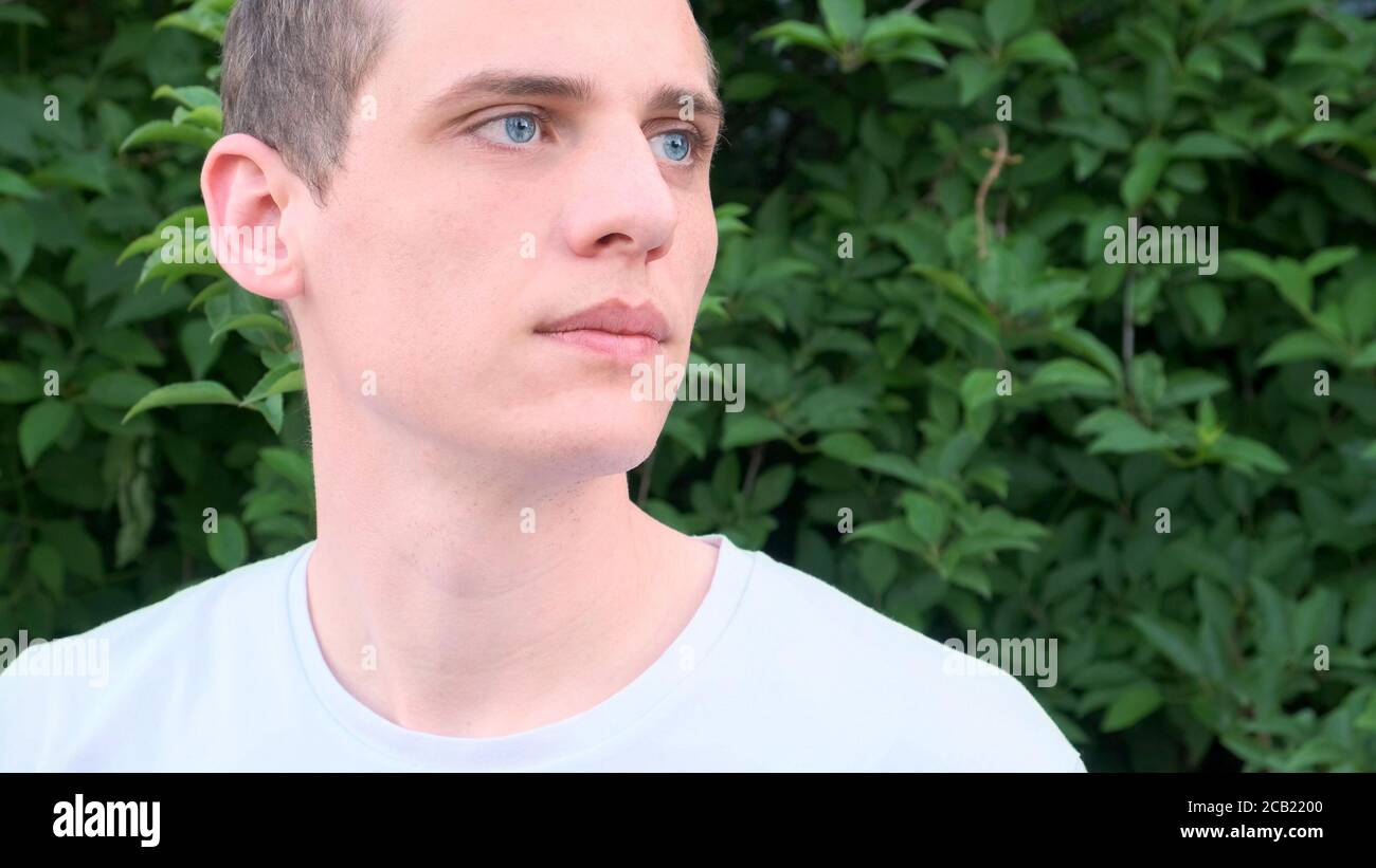 Portrait d'un jeune homme très beau avec des yeux bleus et les cheveux blonds regardant sur le côté contre un arrière-plan de verdure Banque D'Images