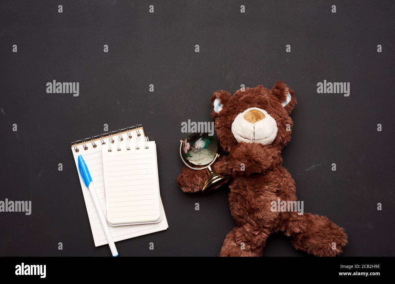 pile de cahiers avec feuilles blanches vierges et un ours en peluche brun tenant un globe de verre sur un fond noir de tableau de craie, de retour à l'école Banque D'Images