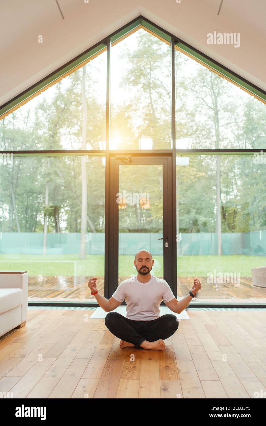 Homme dans des vêtements décontractés assis en position lotus méditer, se détendre. Arrière-plan grande fenêtre panoramique. Yoga à la maison concept Banque D'Images