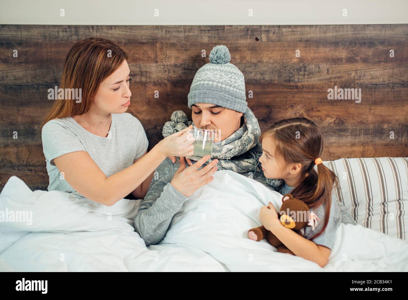 femme soigneuse et petite fille s'assoient sur le lit avec un père malade ayant une température élevée, de la fièvre. femme et enfant le soutiennent et lui donnent des médicaments Banque D'Images