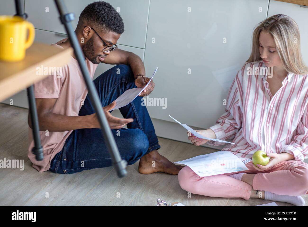 Jeune couple de courses mixtes d'étudiants divers en tenue décontractée, assis ensemble sur le plancher de la cuisine à la maison travaillant à leur cours après s'être concentré Banque D'Images