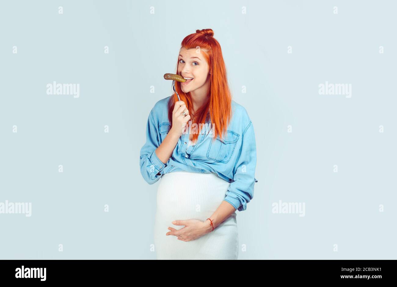 Toujours faim pendant la grossesse. Merveilleuse femme à tête rouge tenant  la main sur le ventre enceinte et piquant délicieux pickle sur la fourche  souriant à la caméra à la lumière Photo