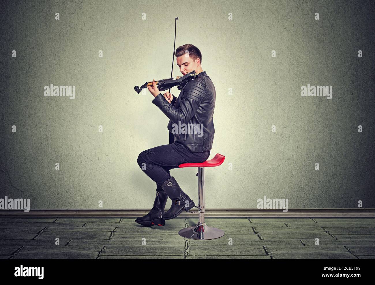 Passion de la musique, concept de passe-temps. Jeune homme habillé moderne et élégant en cuir noir veste jouant sur le violon noir assis sur la chaise rouge dans le studio est Banque D'Images