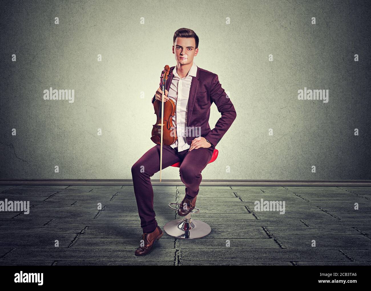 Jeune homme posant tenant un violon moderne et un bâton de violon assis sur un rouge dans la chambre avec un mur et un sol gris. Modèle vertical en bordeaux vineux Banque D'Images