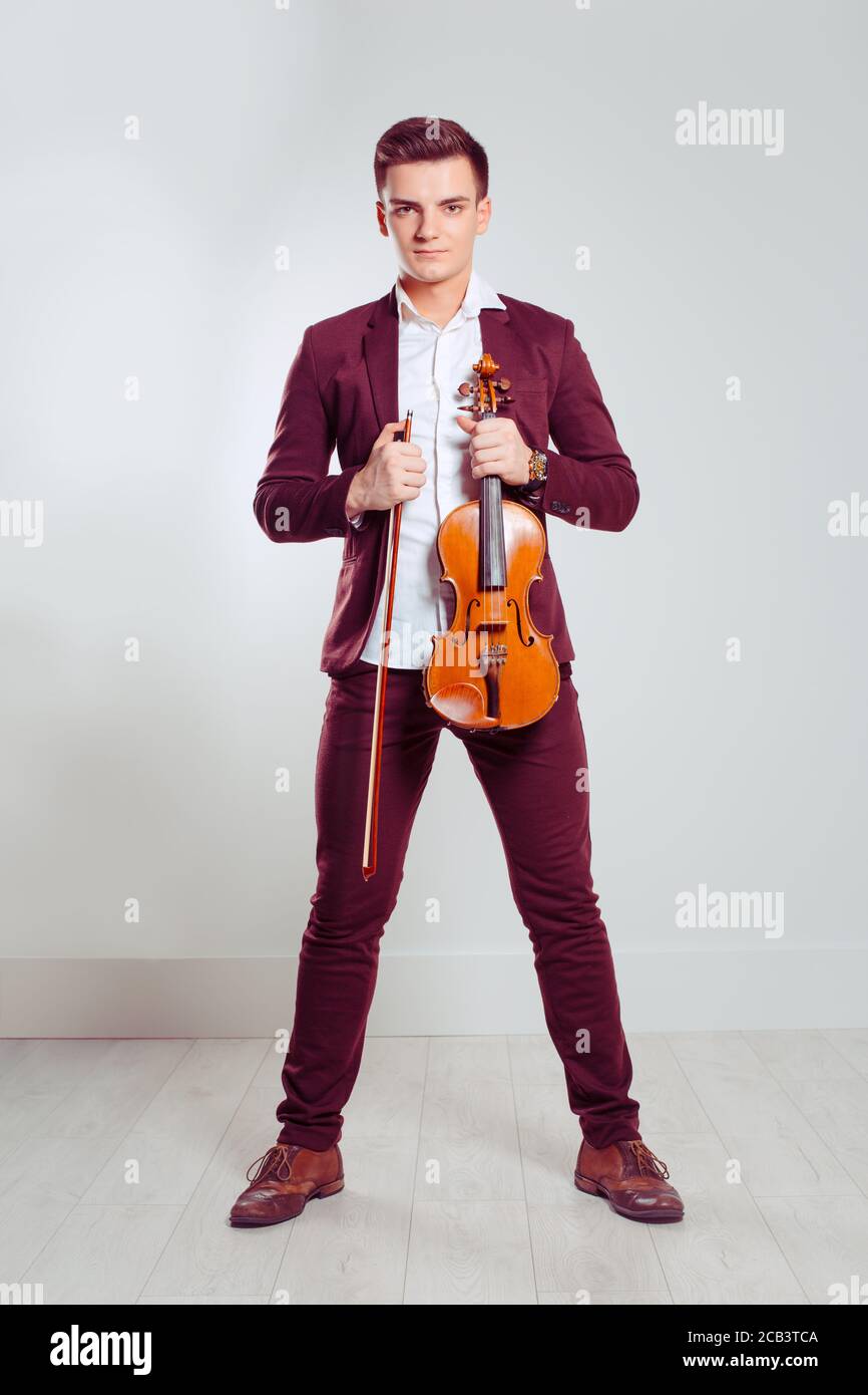 Jeune homme posant tenant un violon moderne et un bâton de violon debout dans une pièce avec un mur et un sol légers. Modèle vertical en costume rouge bordeaux classique Banque D'Images