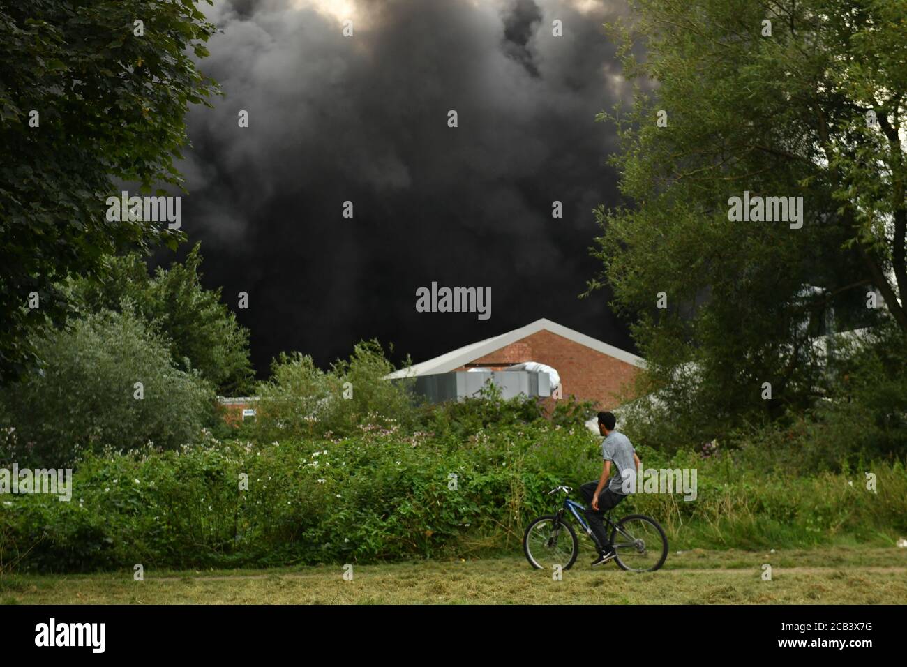 Un homme passe à vélo tandis que la fumée s'illumine d'un feu violent sur un domaine industriel de Birmingham. Les équipes de 10 moteurs d'incendie s'attaquent à l'incendie. Banque D'Images