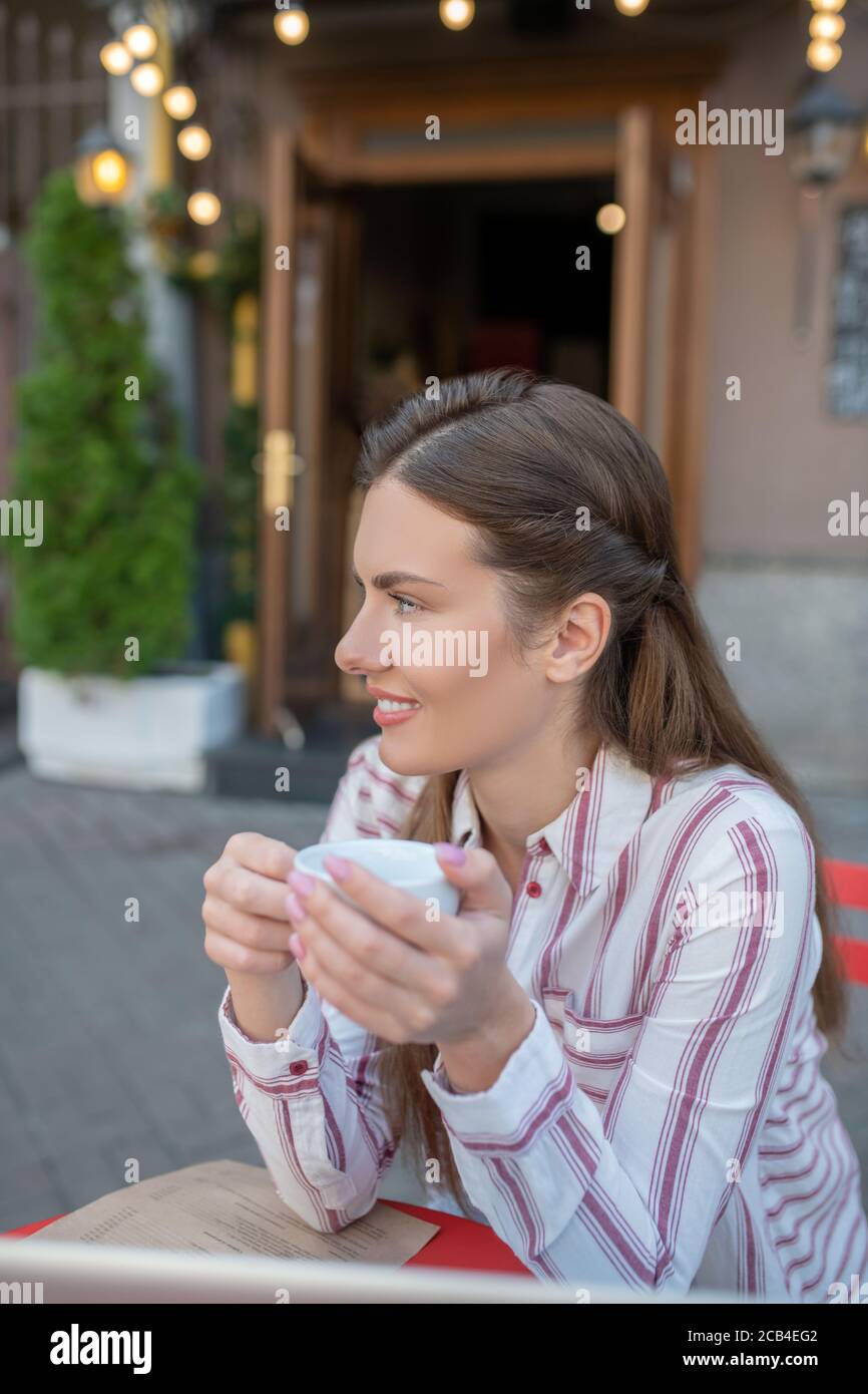 Femme souriante tenant une tasse de café blanc dans un café en plein air, regardant de côté Banque D'Images