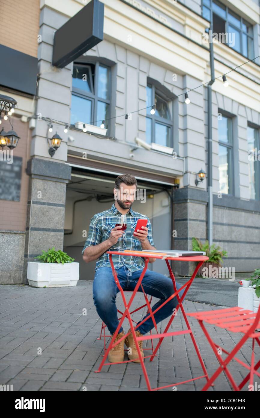 Beau homme barbu du vin rouge, vérifiant son téléphone dans un café en plein air Banque D'Images