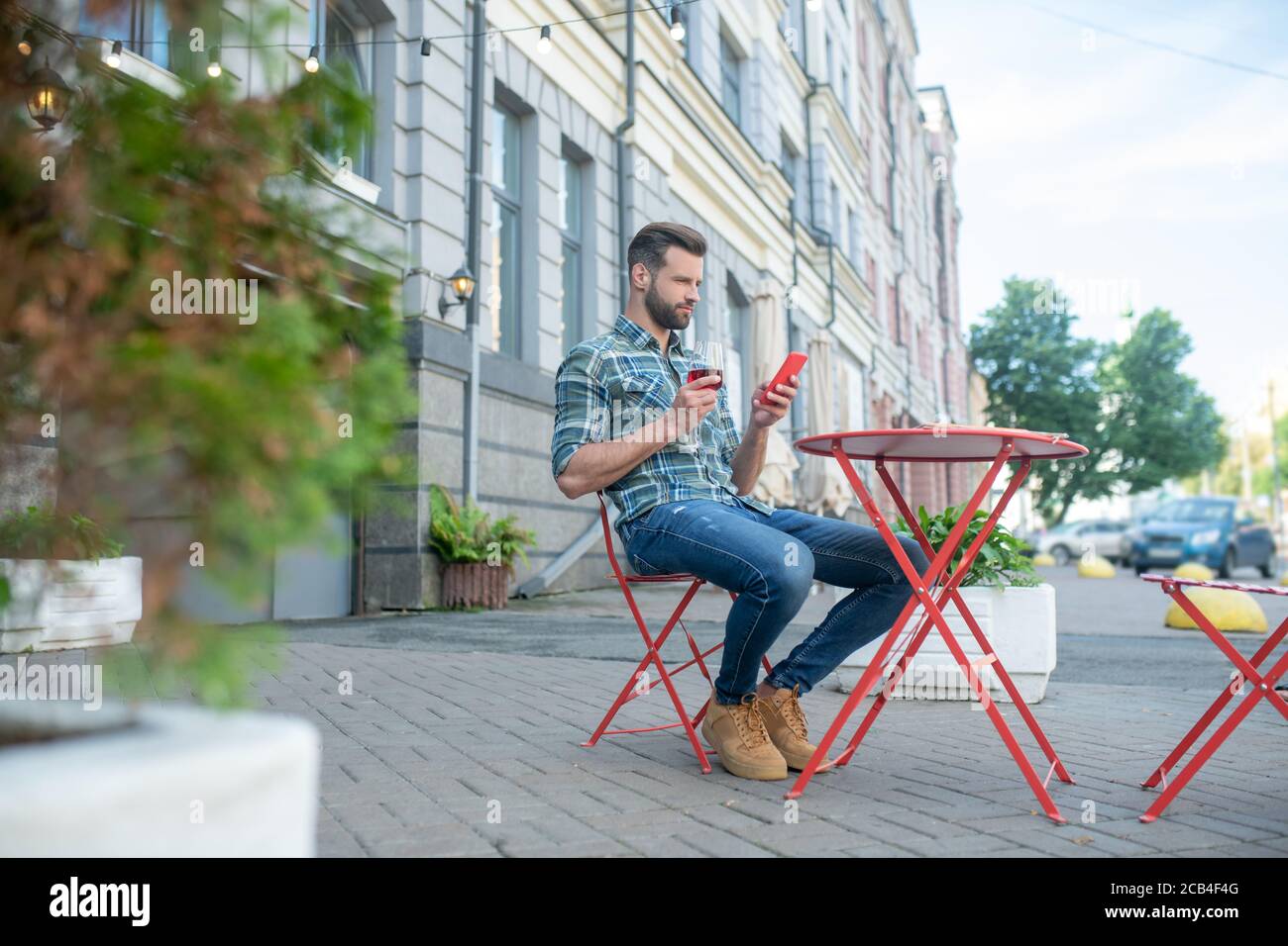 Beau homme à barbe, joyeux verre de vin rouge, en vérifiant son téléphone dans un café en plein air Banque D'Images