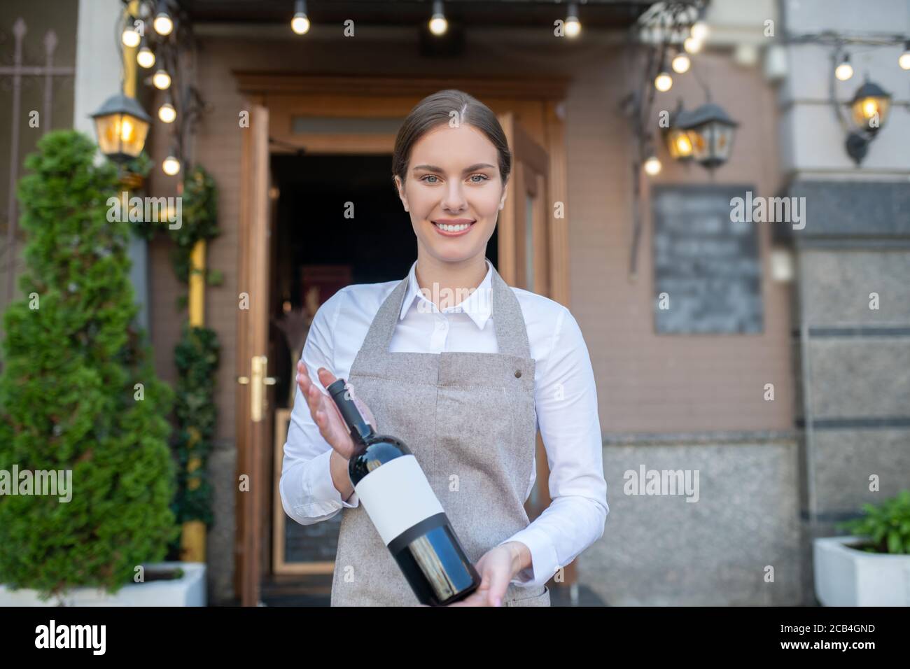 Jolie serveuse souriante sur un tablier gris contenant une bouteille de vin Banque D'Images