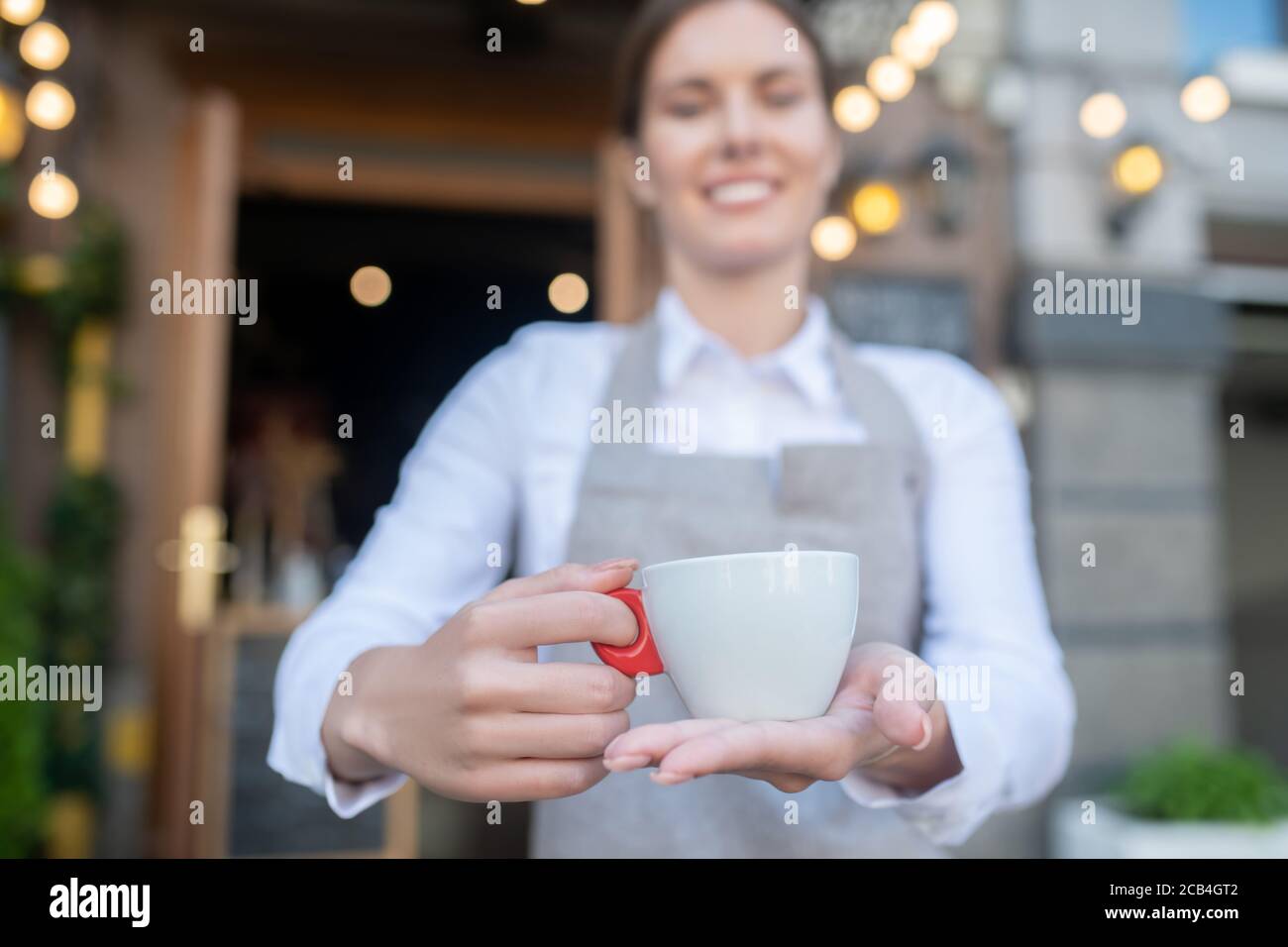 Jolie serveuse en tablier gris offrant une tasse de café Banque D'Images