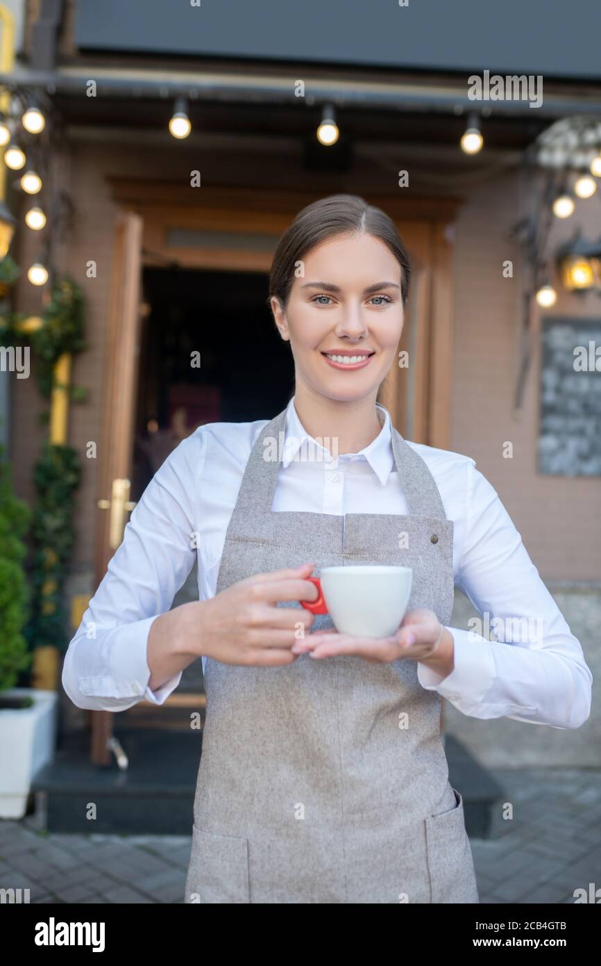 Jolie serveuse souriante sur un tablier gris offrant une tasse de café Banque D'Images