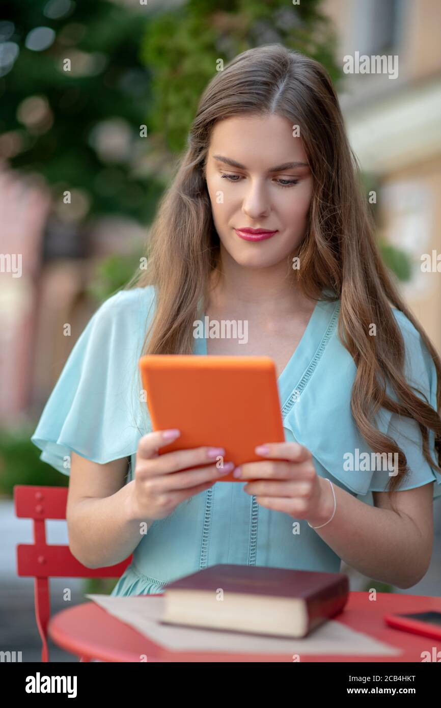 Femme en robe bleue assise dans un café, tenant une tablette orange, regardant l'écran Banque D'Images
