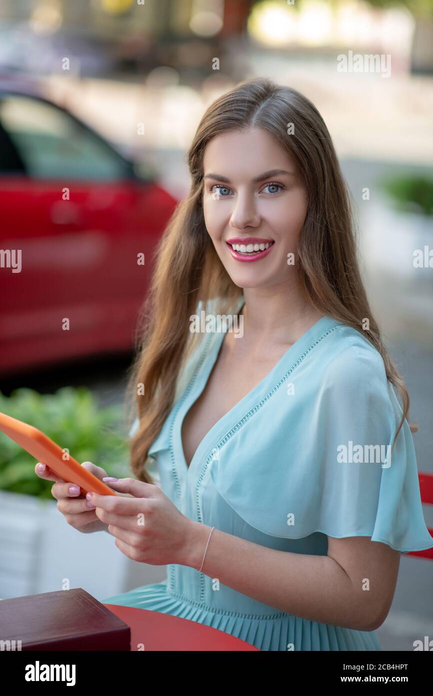 Femme souriante en robe bleue assise dans un café en plein air, tenant une tablette orange Banque D'Images