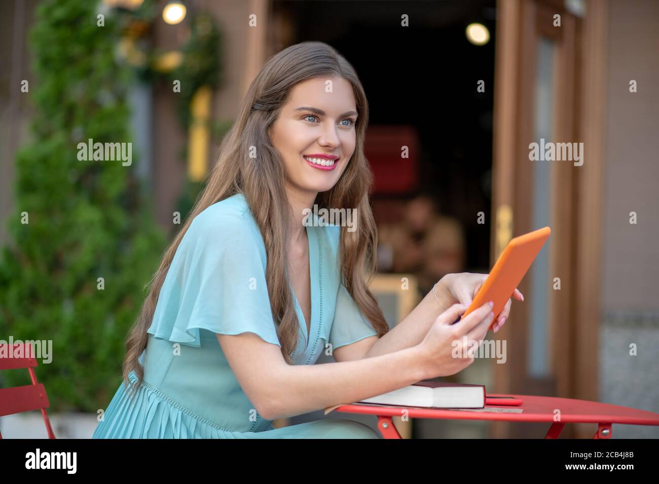 Femme romantique en robe bleue assise dans un café en plein air, tenant une tablette orange, souriant Banque D'Images