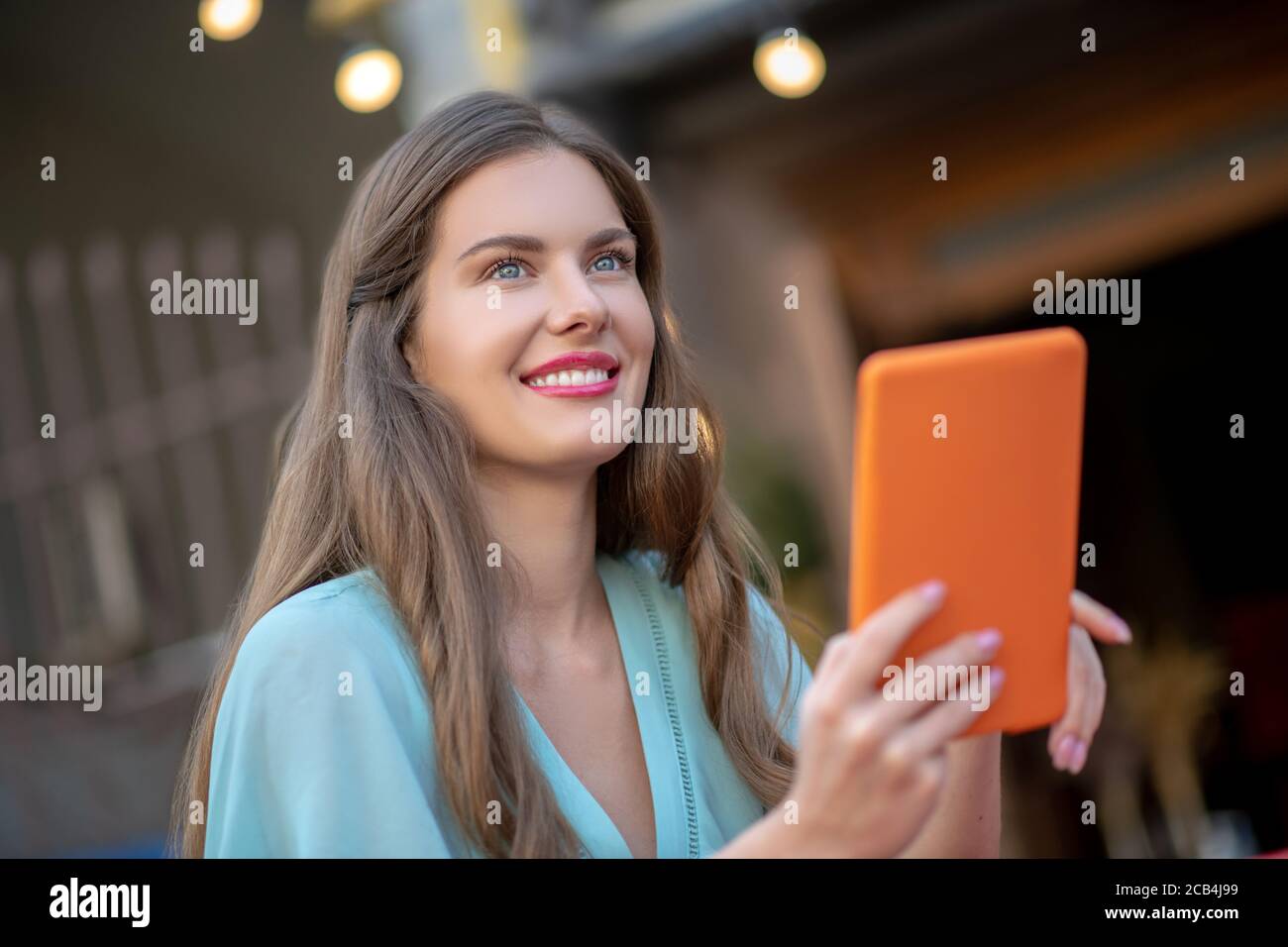Femme romantique en robe bleue assise à l'extérieur, tenant une tablette orange, souriant Banque D'Images
