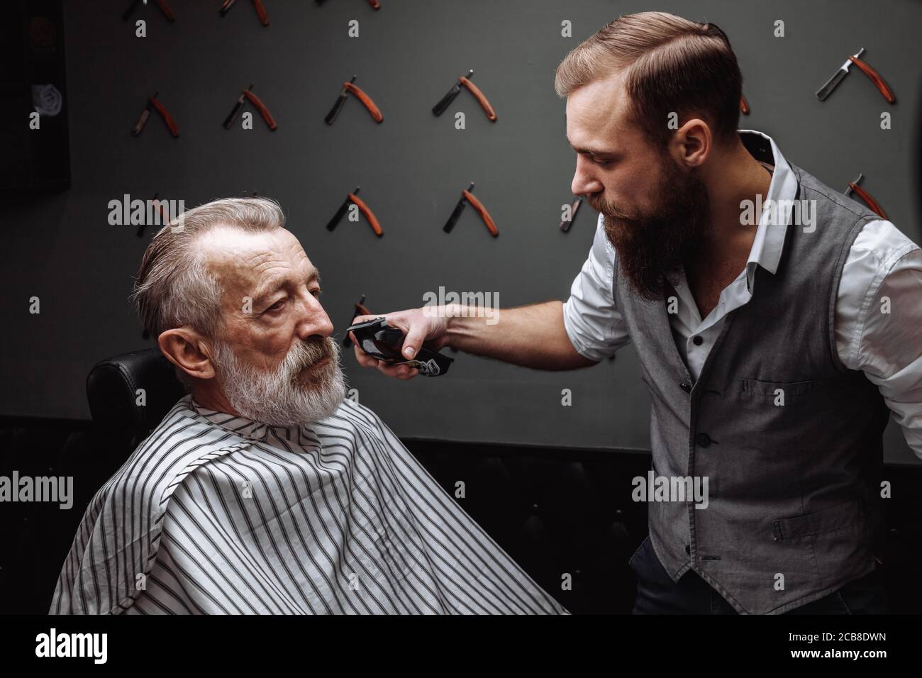 Gros plan d'une coiffure s travailler pour un beau vieux respectable homme barbu d'âge à la coiffure. Il est en train de faire avec le style rasoir électrique. Banque D'Images