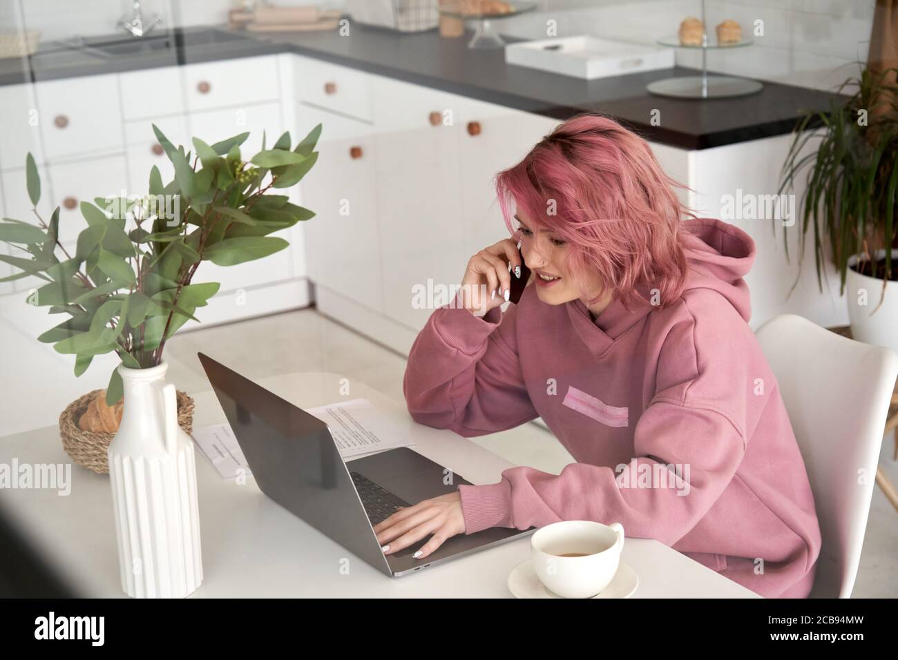 Hipster souriant jeune fille parle au téléphone en utilisant un ordinateur portable assis à la table de bureau à la maison. Banque D'Images