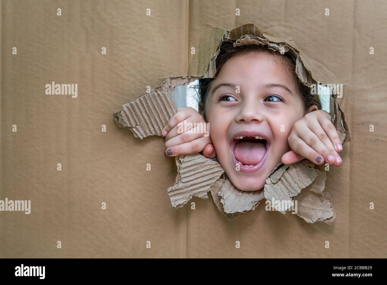 Fille souriante avec sa bouche ouverte derrière un carton. La fille regarde à travers un grand trou dans le carton et avec ses mains sur le bord du h Banque D'Images