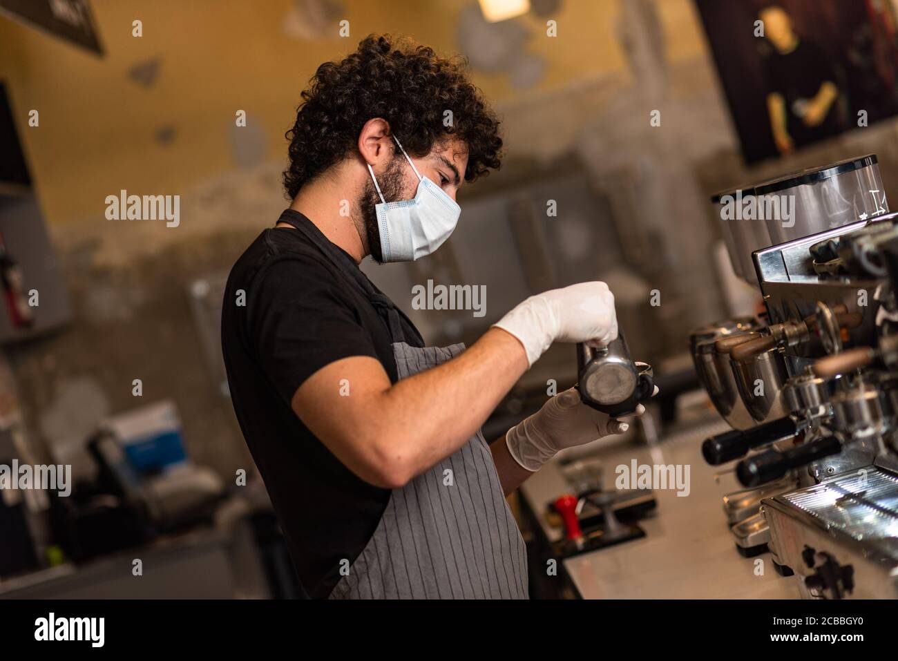 Jeune homme, barista, qui prépare un café expresso tout en portant un masque chirurgical et des gants pour prévenir la propagation du virus corona - concept de travail de sécurité du Bar. Banque D'Images