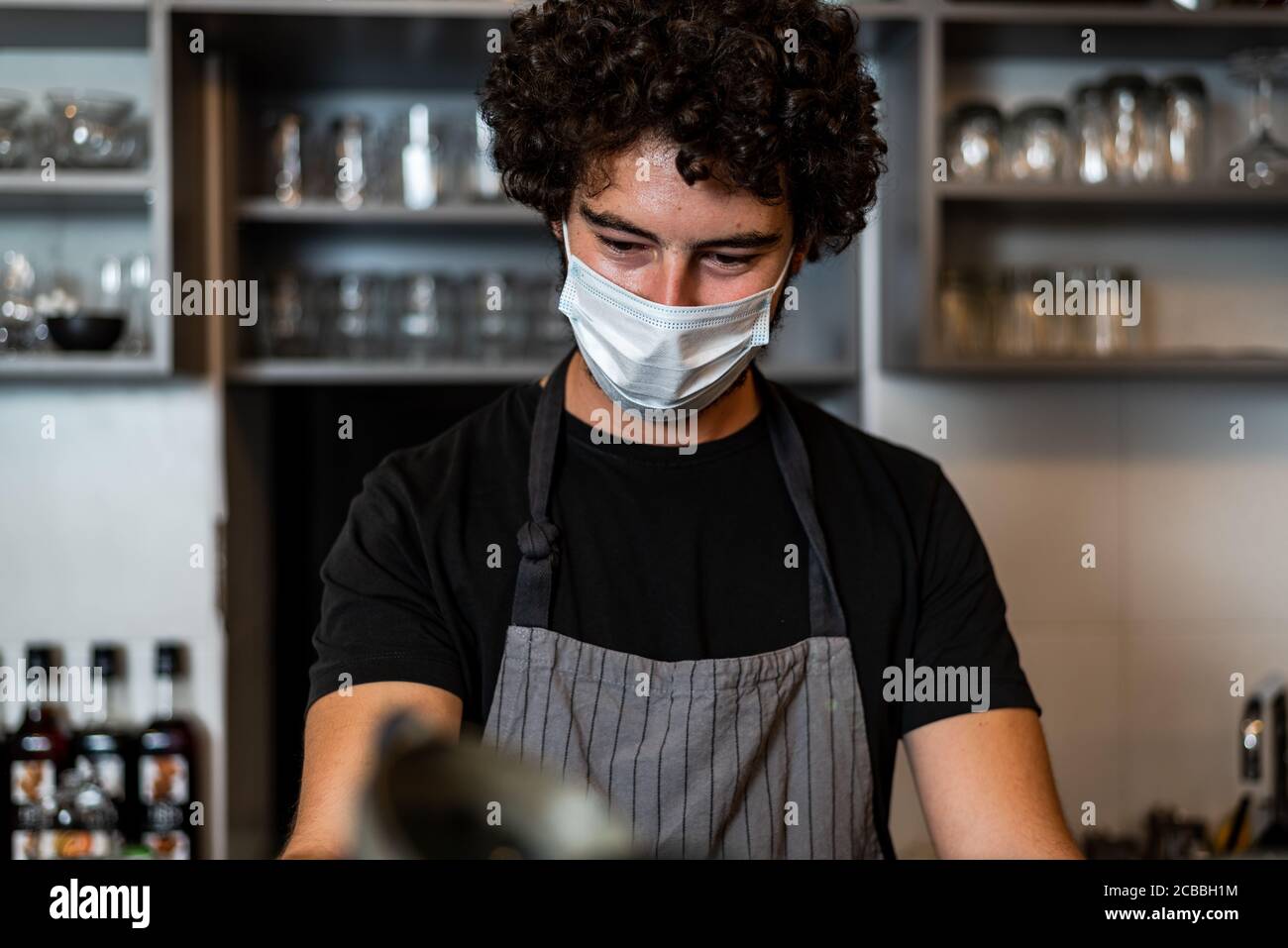 Jeune homme, barista, qui prépare un café expresso tout en portant un masque chirurgical et des gants pour prévenir la propagation du virus corona - concept de travail de sécurité du Bar. Banque D'Images