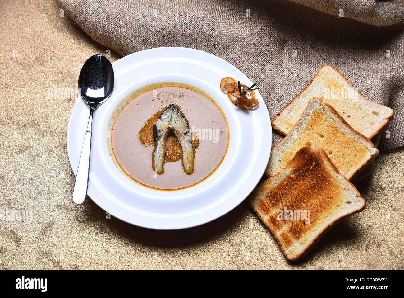 Soupe à la crème ou purée brune sur fond beige. Soupe de poisson avec un morceau de poisson et des rondelles d'oignon frits sur une assiette blanche. Concept de cuisine française. Faites griller les morceaux de pain près de l'assiette sur un chiffon à métaux. Banque D'Images
