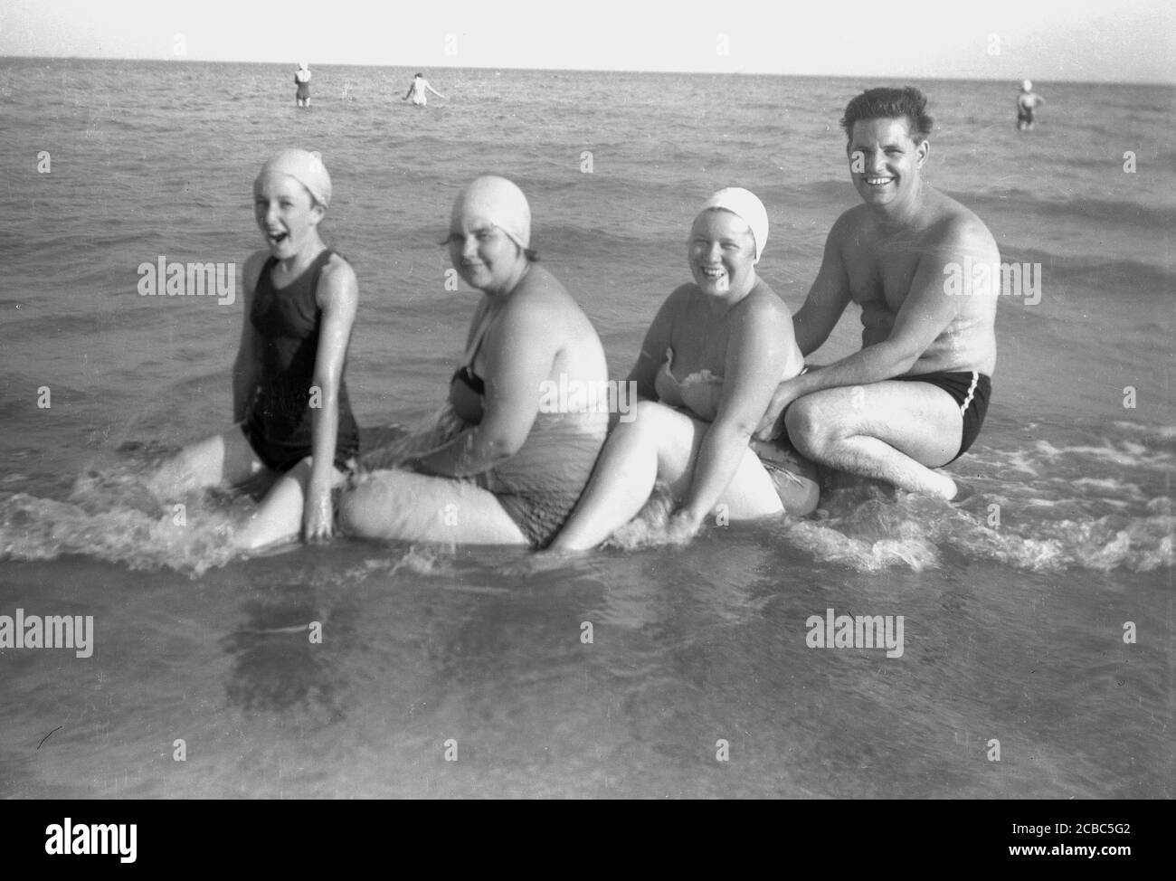 Années 1950, historique, au bord de la mer, une famille s'amusant pendant leurs vacances assis dans les eaux peu profondes de l'océan, posant pour une photo, Angleterre, Royaume-Uni. Banque D'Images