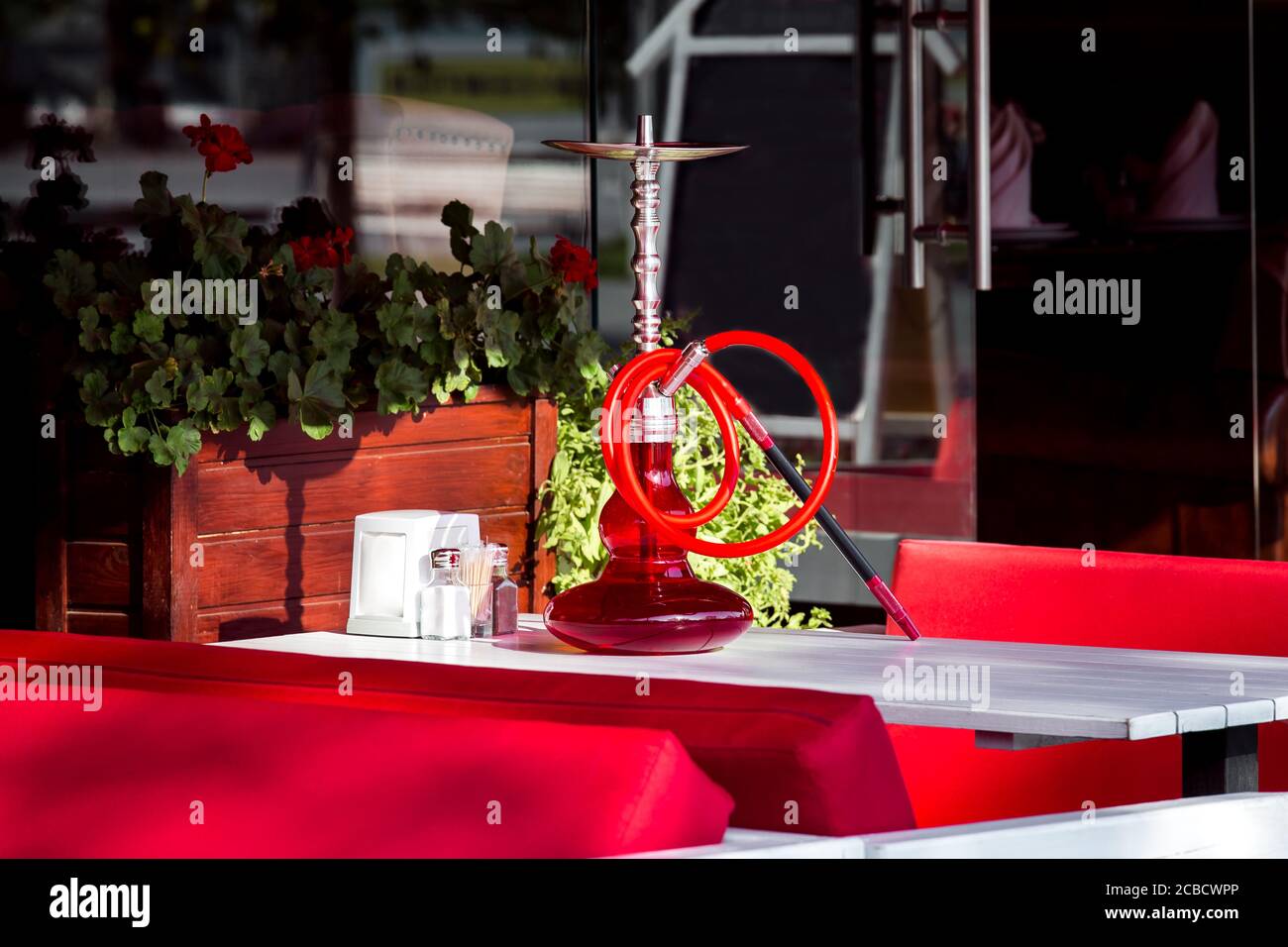 Un narguilé avec une fiole en verre rouge et un tuyau pour fumer sur une table en bois blanc d'un café extérieur avec canapés rouges par temps ensoleillé. Banque D'Images
