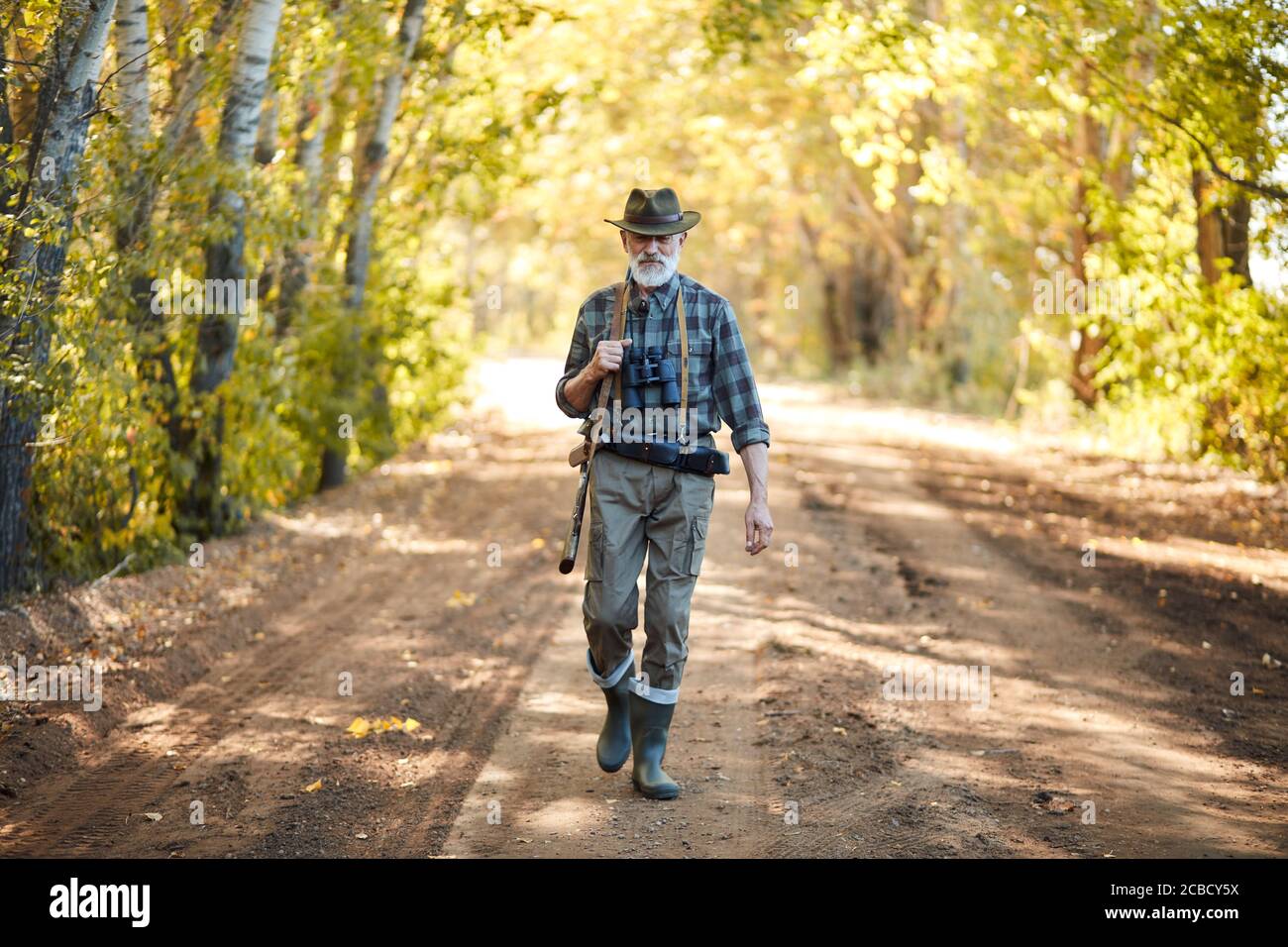 Un homme âgé avec une barbe grise rentre à la maison après avoir chassé dans la forêt, heureux. Porter des bottes en caoutchouc, un chapeau. Tenant le fusil Banque D'Images