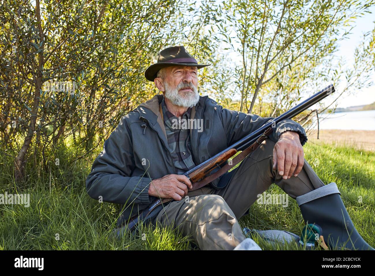 Homme barbu avec fusil de chasse assis sur l'herbe, se reposer après la chasse, porter un chapeau, un manteau et des bottes en caoutchouc Banque D'Images