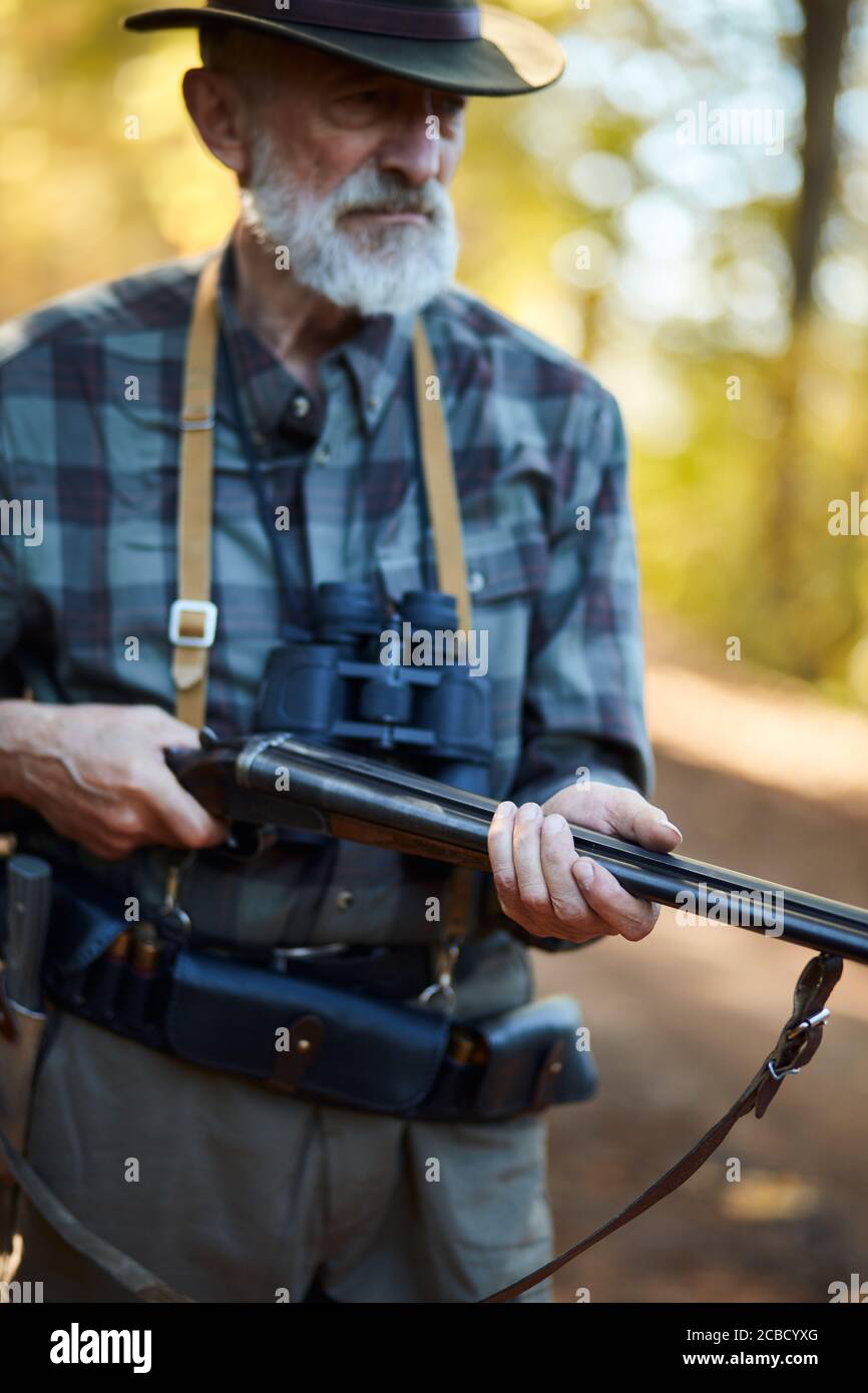 Chasseur plus âgé avec une barbe grise tenant un fusil pour chasser les oiseaux, prêt à tirer. Arrière-plan de la forêt Banque D'Images