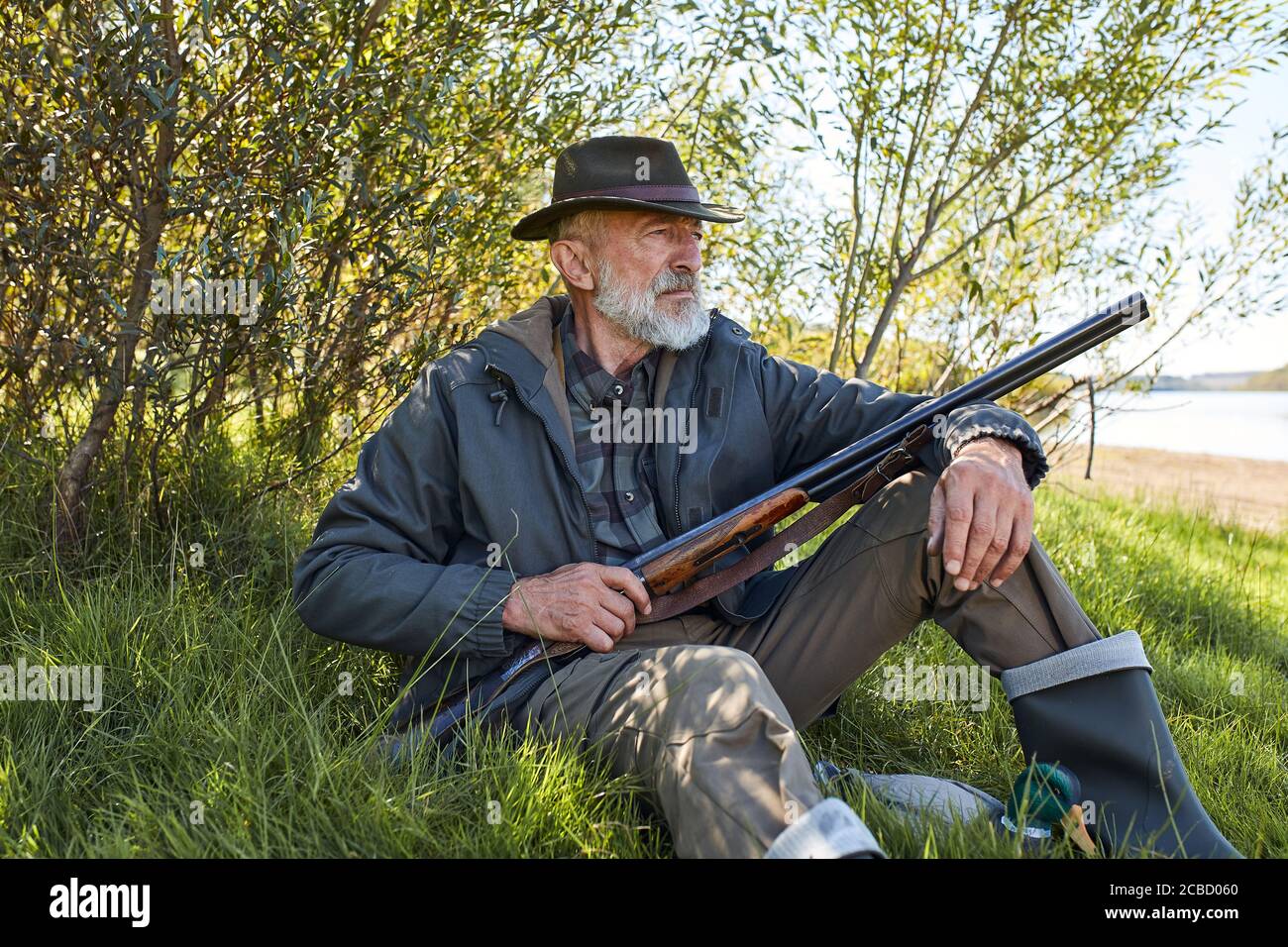 Homme barbu avec fusil de chasse assis sur l'herbe, se reposer après la chasse, porter un chapeau, un manteau et des bottes en caoutchouc Banque D'Images