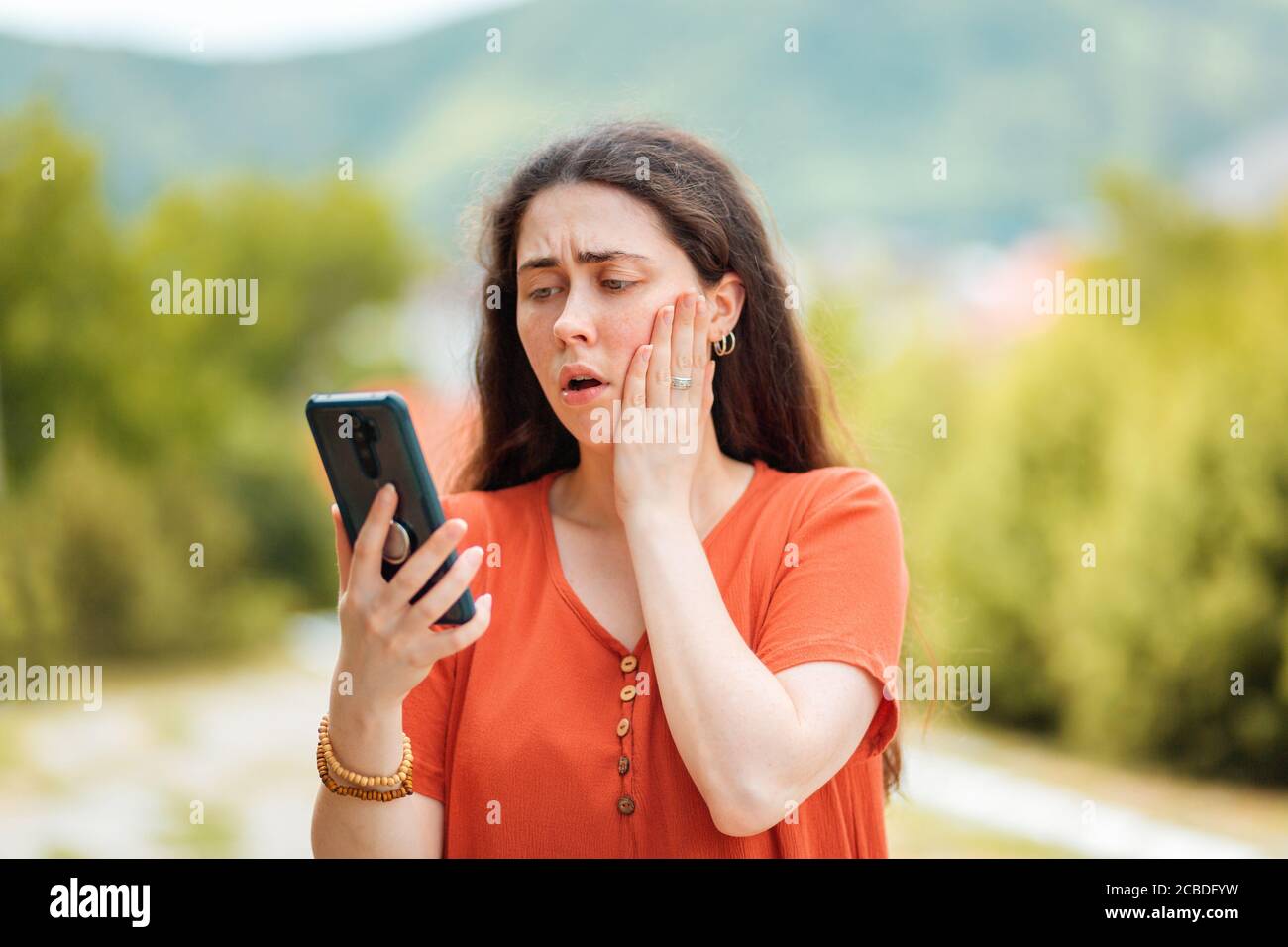 Portrait d'une femme regardant le téléphone, embrassant son visage dans la peur. Le parc est en arrière-plan. Concept de fraude en ligne et de fausses nouvelles. Banque D'Images