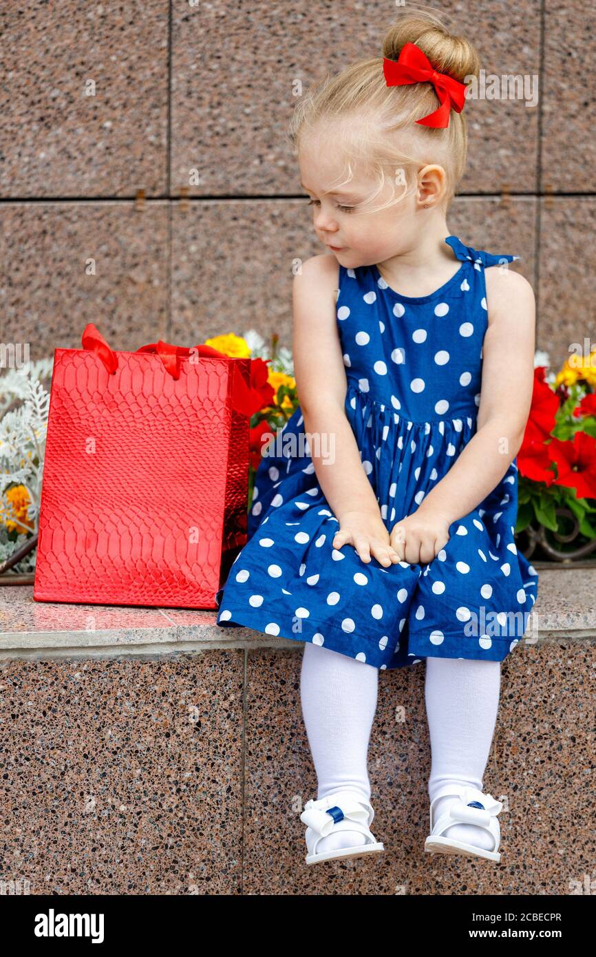 une petite fille dans une robe bleue et une rouge bow est assis avec des  sacs de couleur dans la rue à côté fleurs Photo Stock - Alamy