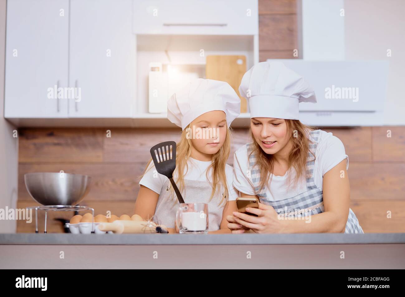 la mère caucasienne et la fille d'enfant en casquettes et tabliers regardent l'écran de smartphone regarder la recette de gâteau délicieuse, allant cuisiner ensemble Banque D'Images