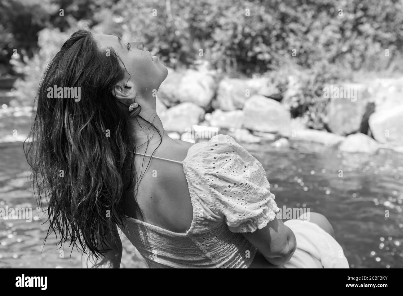 Photo en noir et blanc de derrière une belle jeune femme enceinte aux cheveux longs vêtus de blanc, assise sur les rochers à côté d'une rivière. Banque D'Images